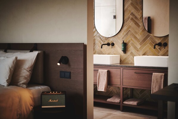 Un dormitorio moderno con cama, mesita de noche y un amplificador Marshall. Junto a él, un baño con salpicadero de azulejos tipo espiga, dos lavabos y un espejo.