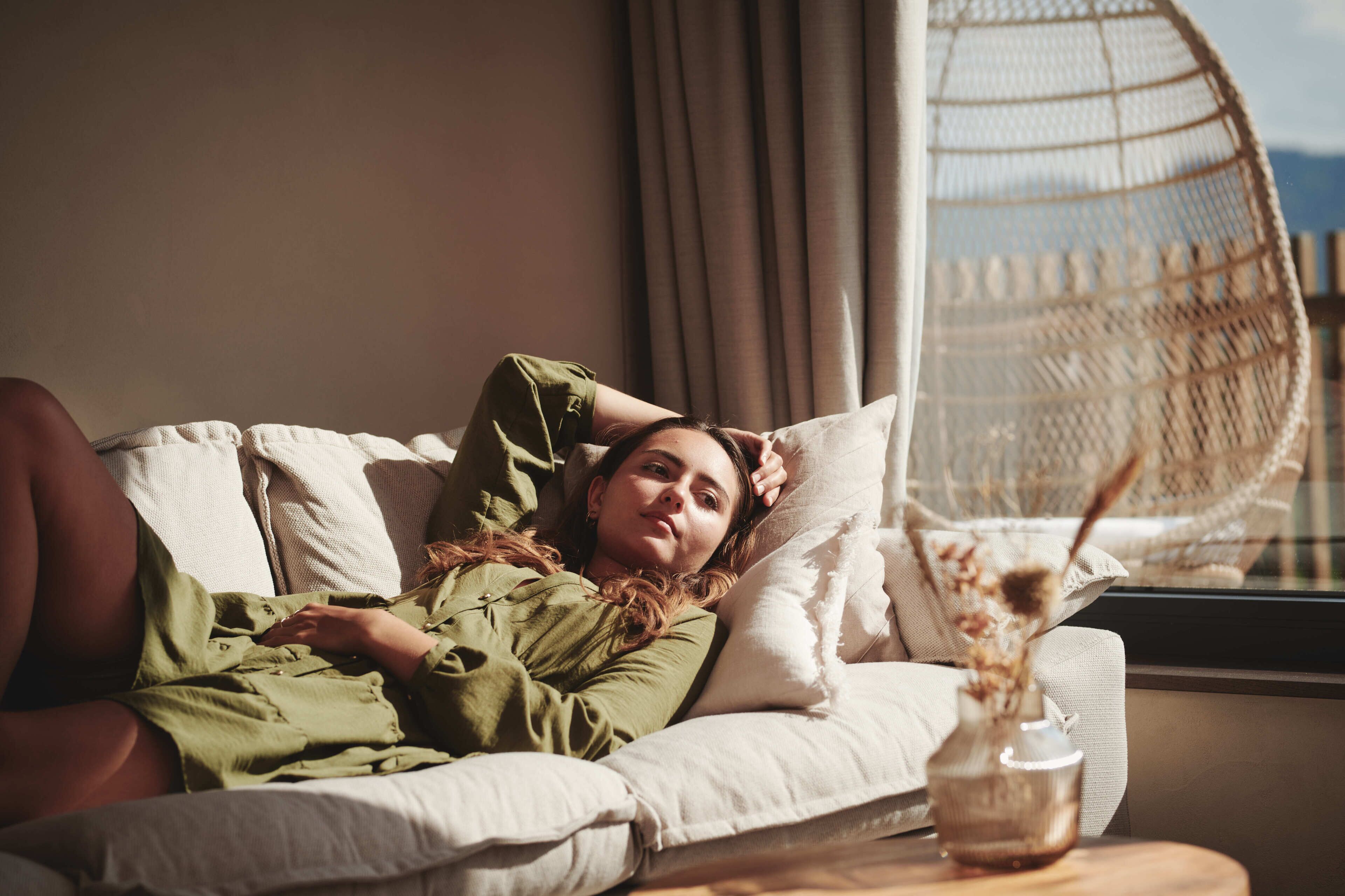 A person lying on a sofa in a room with a wicker chair and a vase with dried flowers on a side table.