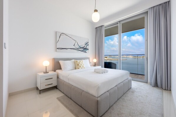 A modern bedroom with a large bed, a nightstand, a bedside lamp, and a view of the sea through a sliding glass door.