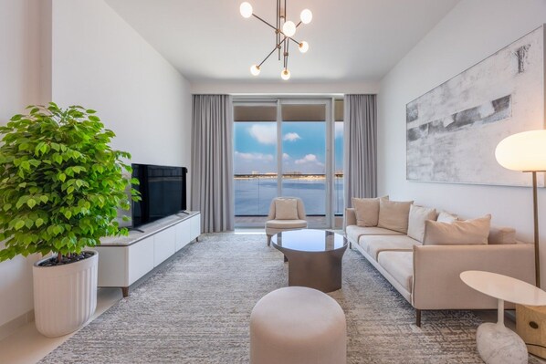 Een moderne woonkamer met een flatscreen televisie, een grote potplant, een bank, een salontafel en uitzicht op zee door grote ramen.