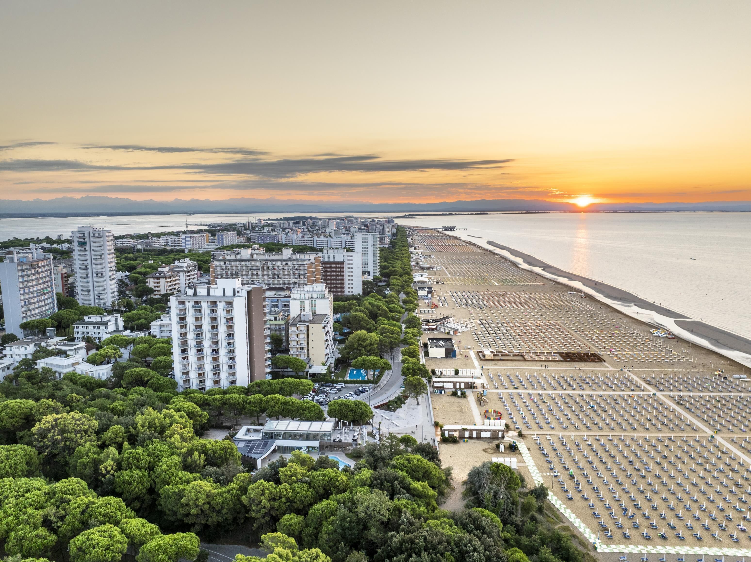 Ilmakuva rannikkokaupungista, jossa näkyy korkeita rakennuksia, aurinkovarjoilla varustettu ranta ja auringonlasku taustalla.