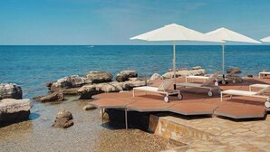 A beachside lounge area with sun loungers and umbrellas, set against a backdrop of the sea and rocky shore. - Vacation complex Istrian Villas Villa V SV2P2 for 4 persons (Umag)