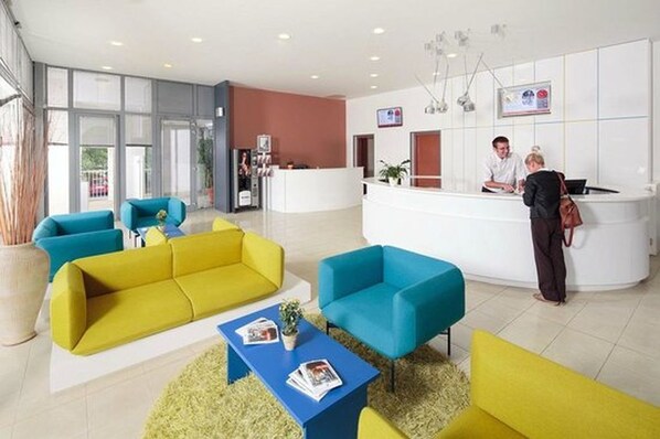 A modern hotel lobby with a reception desk, bright yellow and blue sofas, and a large window providing natural light.