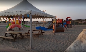 Una playa con una carpa, mesas de picnic y juegos infantiles.