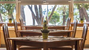 A dining area with wooden tables and chairs, potted plants, and a view of trees through large windows.