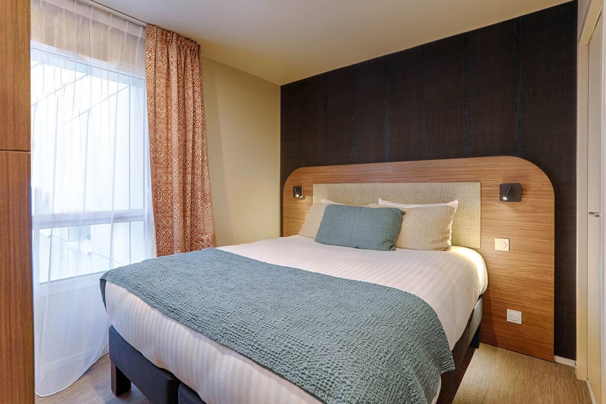 A neatly made bed with a wooden headboard, a patterned curtain, and a window with sheer curtains.