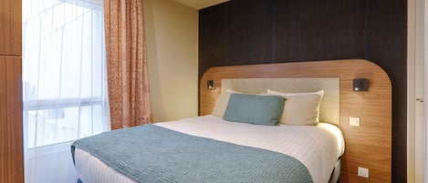 Un lit bien fait, avec une tête de lit en bois, un rideau à motifs et une fenêtre ornée de rideaux légers.
