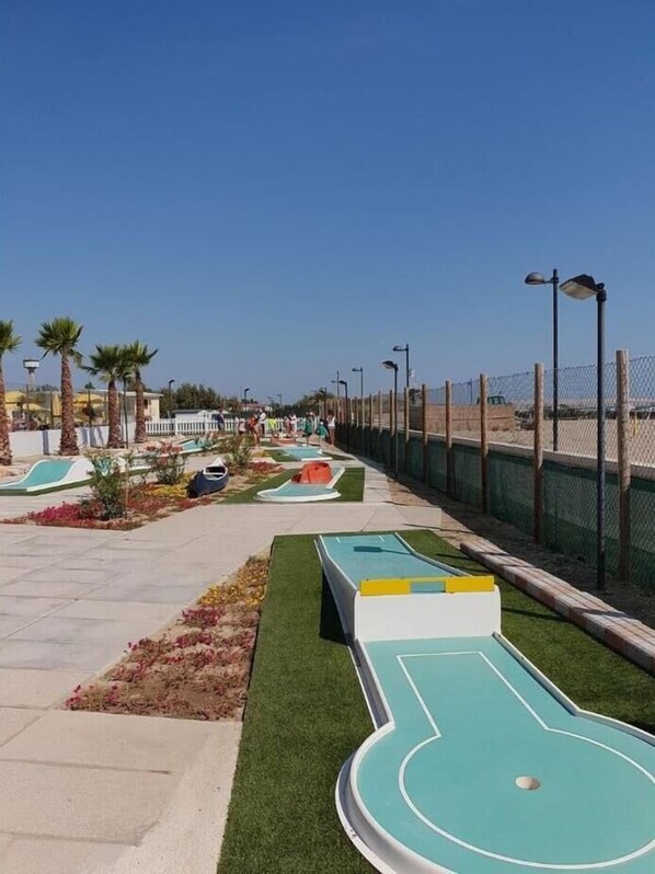 A sunny outdoor area with shuffleboard tables, palm trees, and a fenced walkway. - Vacation park Rosapineta Sud Mobilehome C6 for 6 persons (Rosolina Mare)