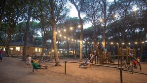 Un parque infantil con columpios, toboganes y una estructura para escalar, rodeado de árboles y luces colgantes.