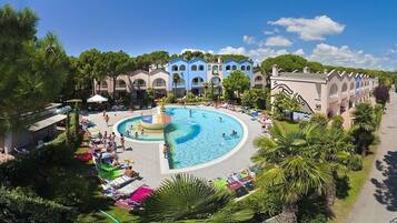 Ein Resort mit einem zentral gelegenen Swimmingpool, umgeben von Palmen und einem Gebäude mit blau-beiger Architektur.