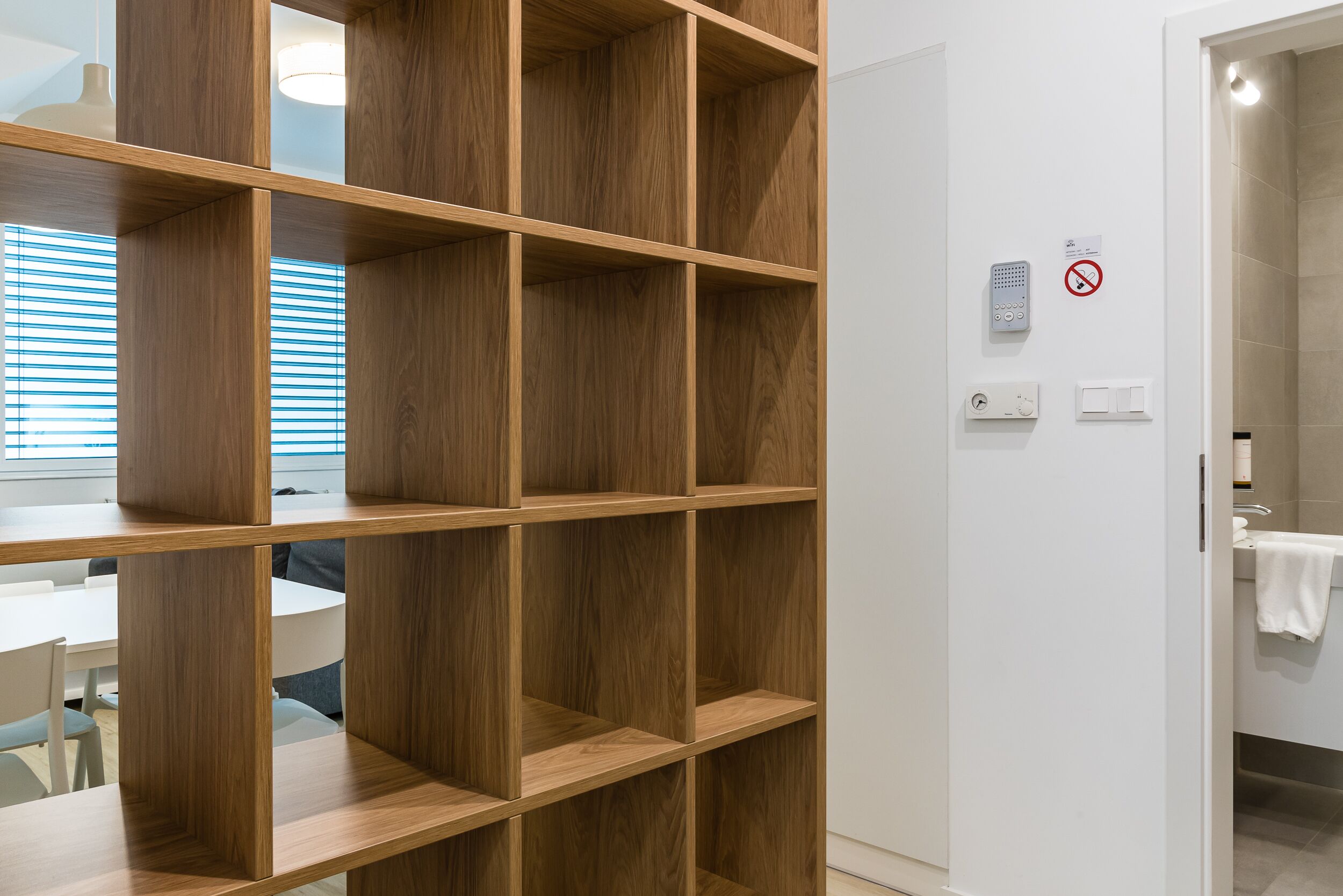 A modern room with a wooden shelving unit, a white wall with a no-smoking sign, and a bathroom door.