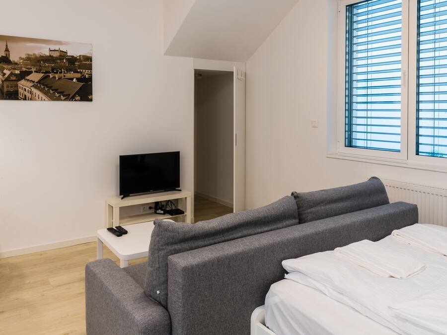 A modern hotel room with a grey sofa set, a flat-screen TV on a white stand, a bed with white linens, and a cityscape painting on the wall.