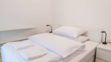 A neatly made bed with white linens and pillows, a bedside table with a lamp, and a white nightstand.