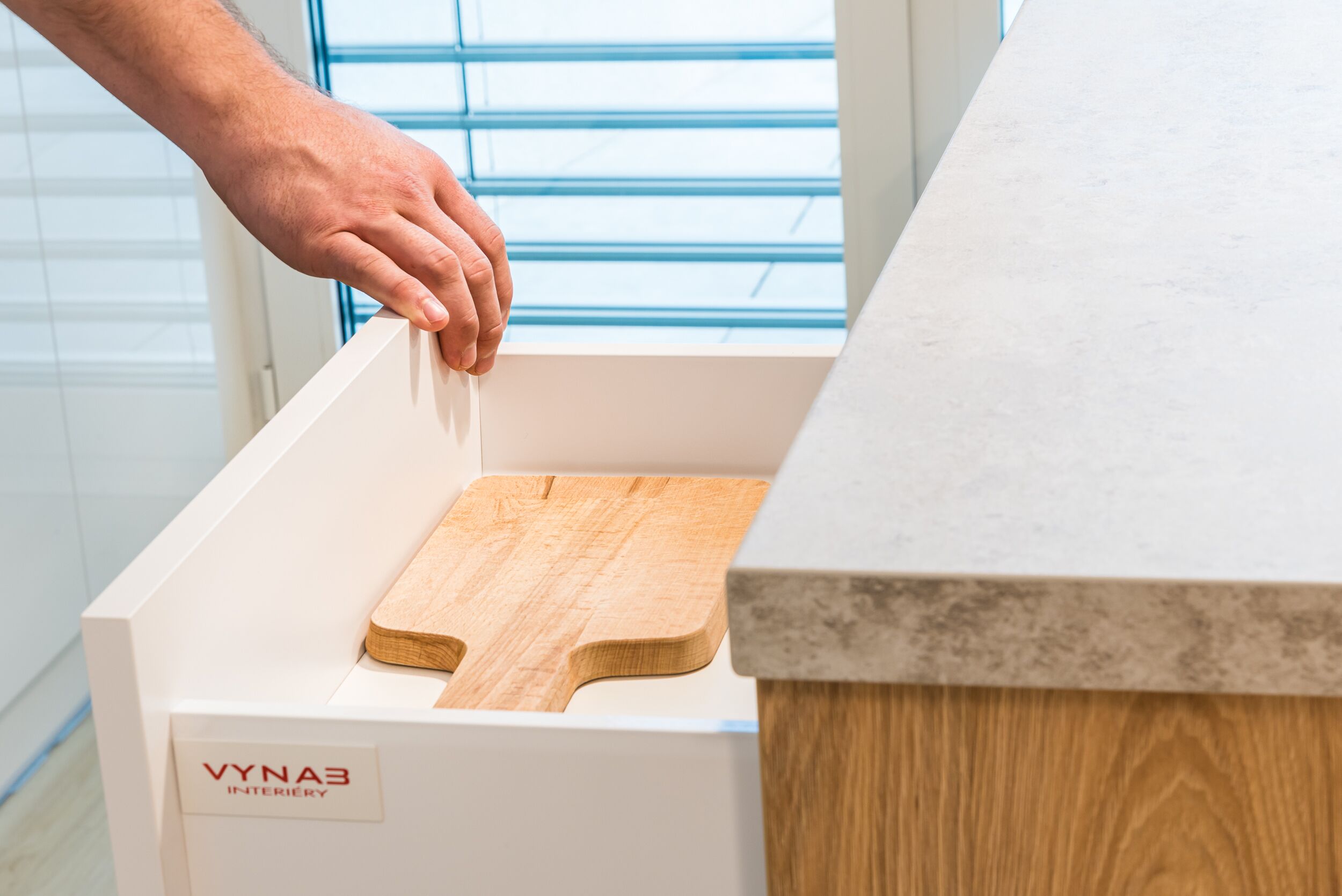 A kitchen with a white cabinet, a wooden cutting board, and a window with blinds.