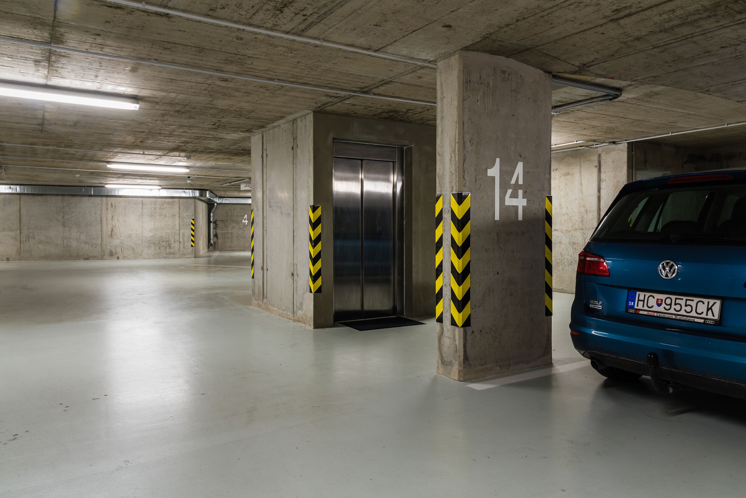Een parkeergarage met betonnen pilaren, een lift en een blauwe auto geparkeerd nabij paal nummer 14.