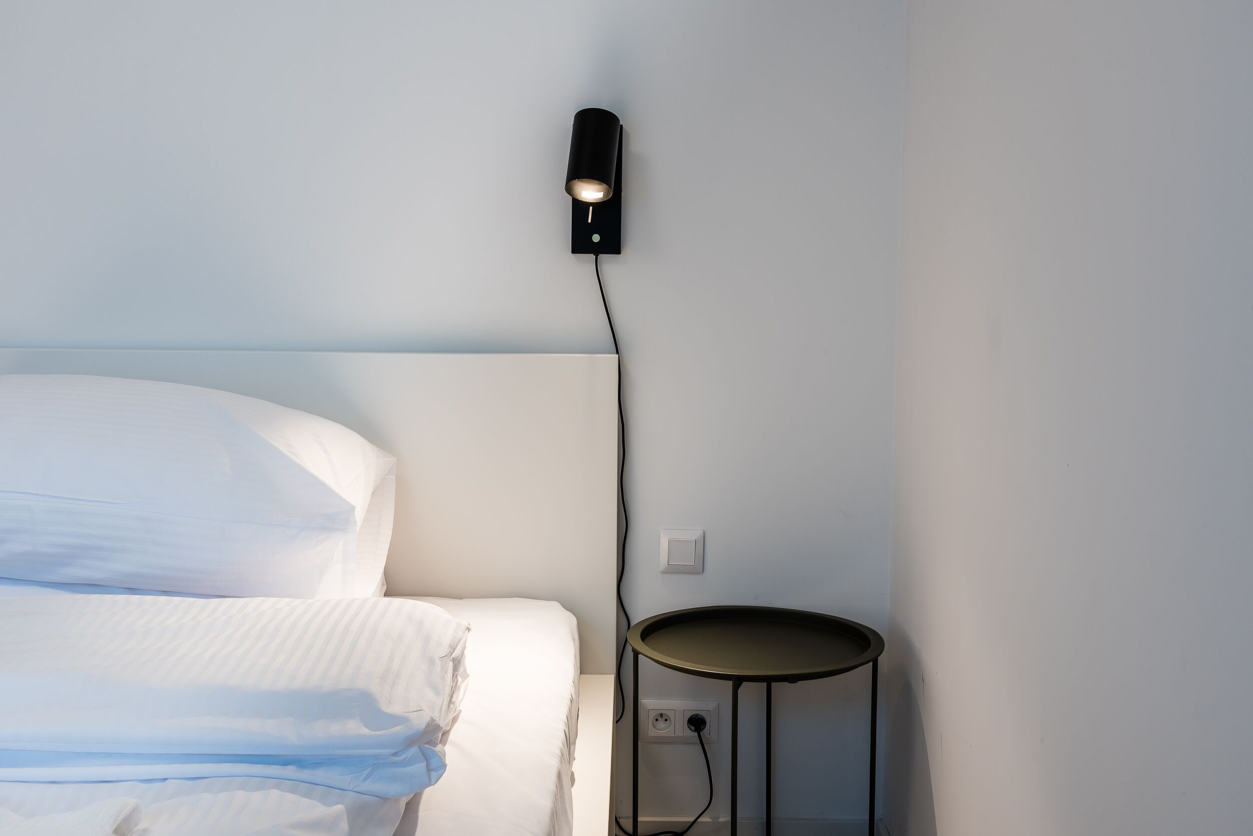 Un lit bien fait, avec du linge de lit blanc, une petite table ronde et un luminaire fixé au mur.
