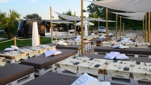 Outdoor lounge chairs with tables, arranged in rows, under a canopy.