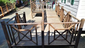 Une terrasse en bois dotée d’une rambarde, qui mène à un espace de détente abrité, aménag é avec des tables et des chaises.