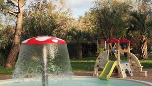 Une fontaine à eau surmontée d’une sorte de champignon, une aire de jeux avec un toboggan et des arbres en arrière-plan.