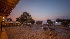 An outdoor patio with stone flooring, tables, and chairs, surrounded by trees and a view of the sea.