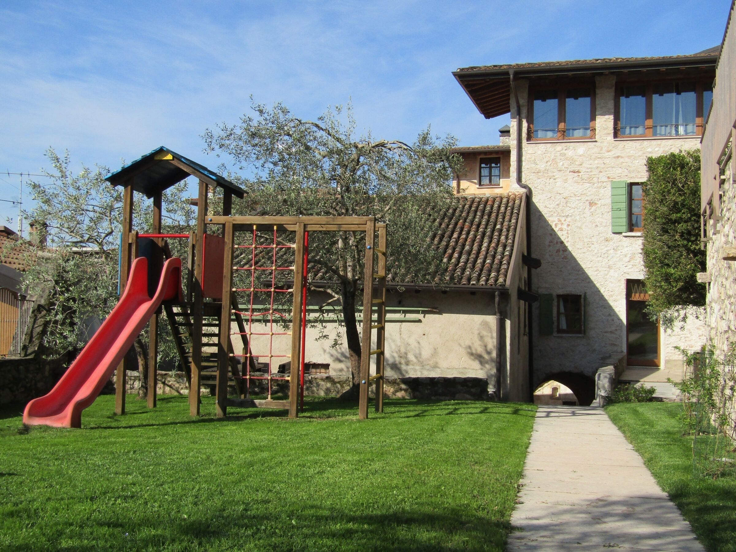 Un terrain de jeux avec un toboggan rouge et une structure d’escalade, un sentier menant à un bâtiment en pierre doté d’un toit en tuiles et de volets verts.