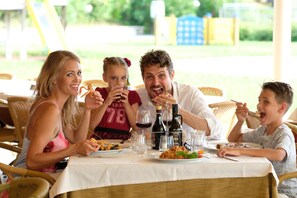 A family dining at an outdoor table with a white tablecloth, surrounded by wicker chairs, in a bright, sunny setting.