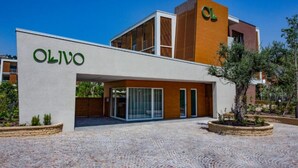 Ein modernes Gebäude mit der Aufschrift "OLIVO" an der Fassade, das Glas- und Holzelemente kombiniert, und ein gepflasterter Innenhof mit Pflanzgefäßen.