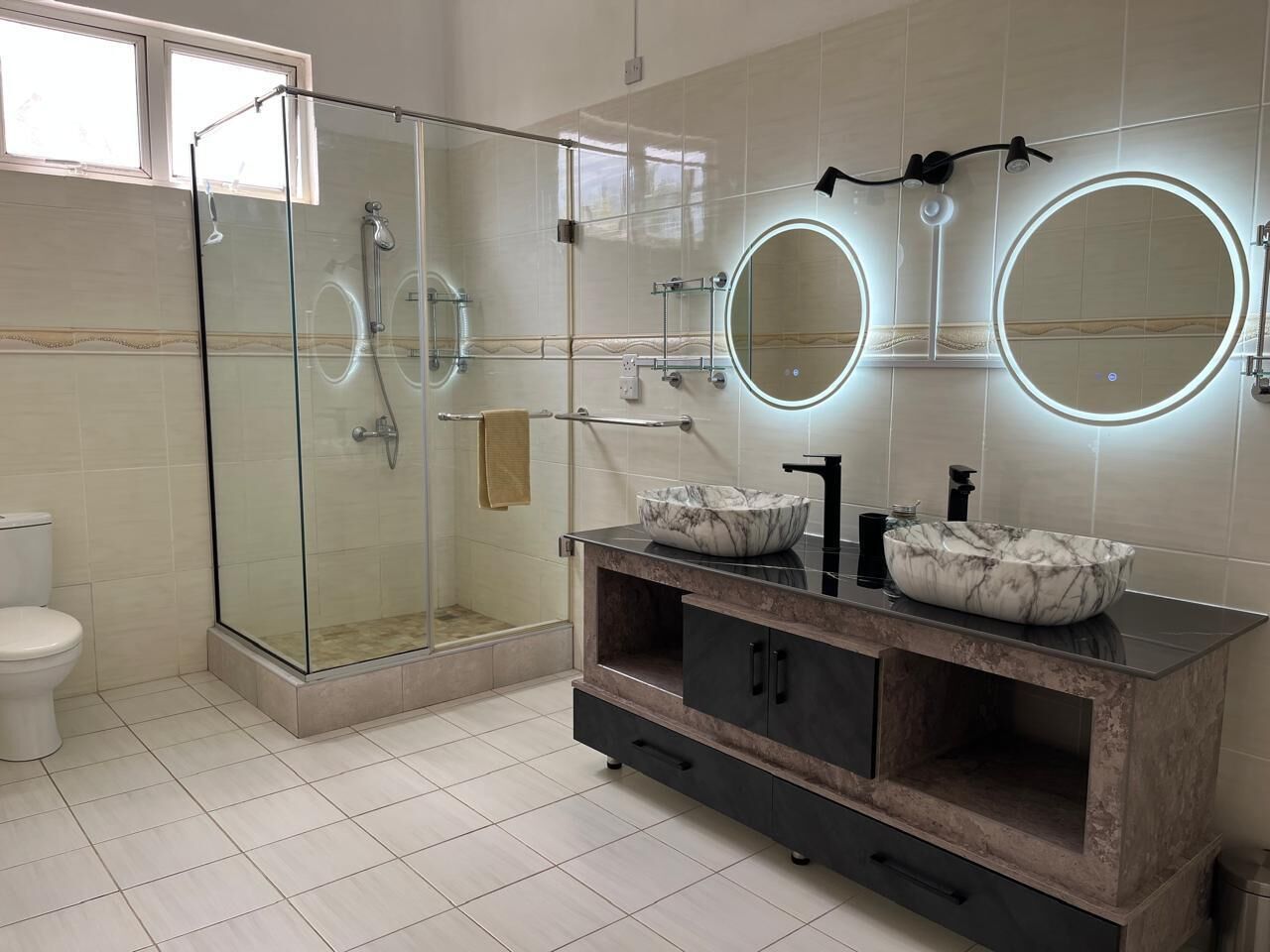 Une salle de bain moderne avec une douche à parois de verre, deux miroirs ronds aux bords éclairés, un lavabo en marbre et un meuble-lavabo en bois foncé.