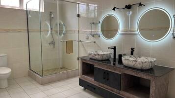Une salle de bain moderne avec une douche à parois de verre, deux miroirs ronds aux bords éclairés, un lavabo en marbre et un meuble-lavabo en bois foncé.
