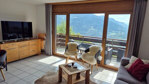 Un salon équipé d’un téléviseur, d’un canapé, d’une table basse et offrant une vue sur les montagnes grâce à de grandes fenêtres.