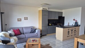 Un salon moderne avec un canapé gris, une petite table basse en bois et un fauteuil gris agrémenté d’un plaid moelleux. On aperçoit, au fond, une cuisine avec des meubles gris et un plan de travail noir.
