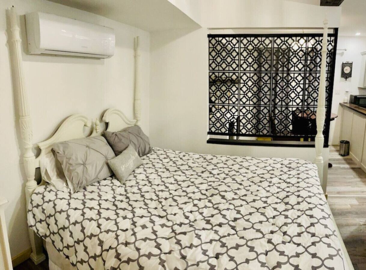 A bedroom with a bed, pillows, an air conditioning unit, a window with a decorative screen, and a small kitchenette area.