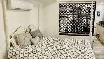 A bedroom with a bed, pillows, an air conditioning unit, a window with a decorative screen, and a small kitchenette area.