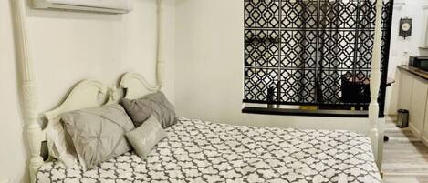 Un dormitorio con cama, almohadas, equipo de aire acondicionado, ventana con celosía decorativa y una pequeña zona de kitchenette.