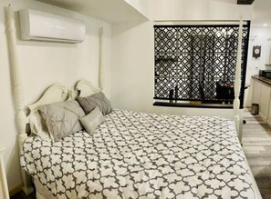Un dormitorio con cama, almohadas, equipo de aire acondicionado, ventana con celosía decorativa y una pequeña zona de kitchenette.