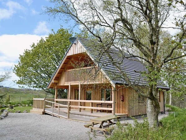 A wooden cabin with a balcony, surrounded by trees and a gravel path. - Deveron Valley Cottages (Huntly)
