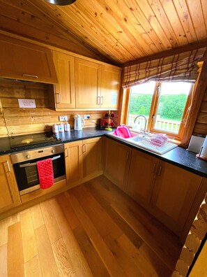 A kitchen with wooden cabinets, a stainless steel oven, a sink with a red dish rack, and a window overlooking a deck. - Deveron Valley Cottages (Huntly)