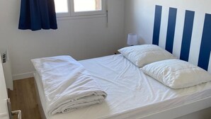 A neatly made bed with white linens and pillows, a window with curtains, and a wall-mounted air conditioner. - Appartement T2 Refait à Neuf (Cabourg)
