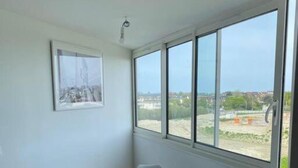 A small room with a white table and chairs, a bench, and a large window offering a view of a construction site. - Appartement T2 Refait à Neuf (Cabourg)