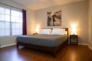 A bedroom with a large bed, two bedside tables with lamps, a window with blinds, and a painting on the wall. - Cozy and Spacious Denton Getaway (Denton)