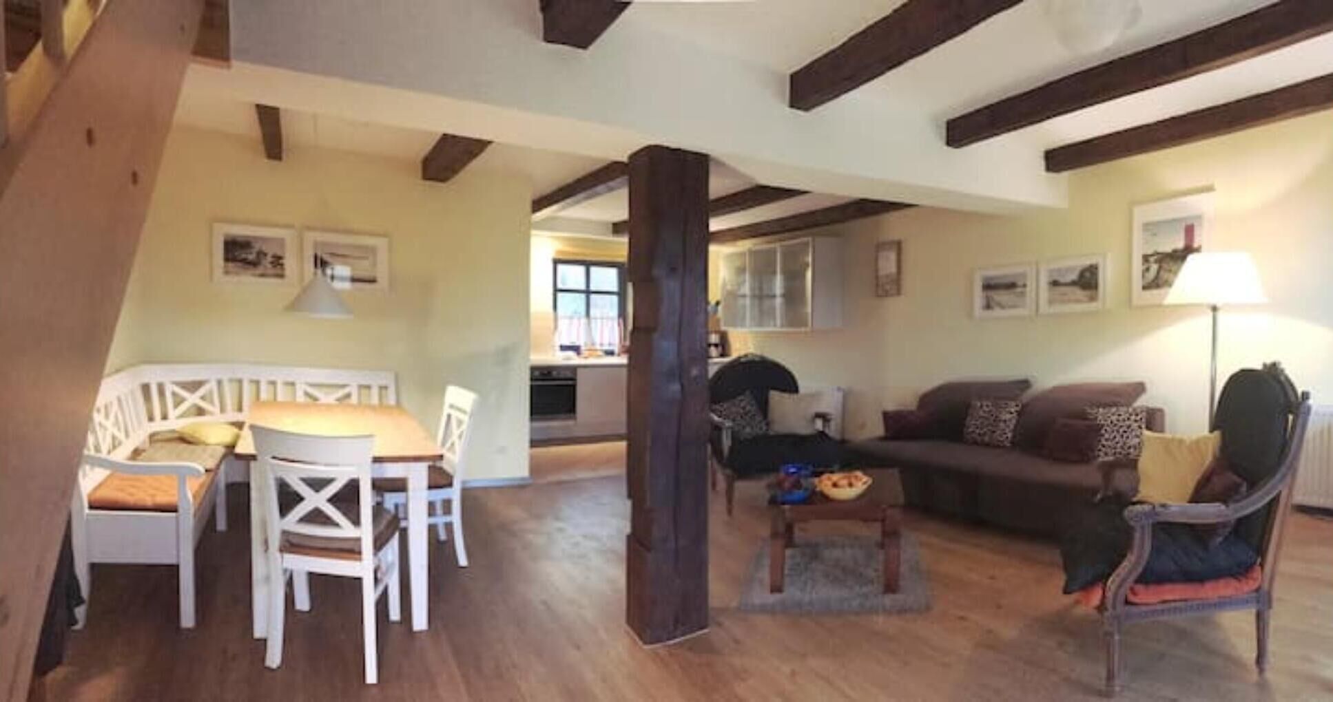 Una sala acogedora con suelo de madera, una mesa de comedor con sillas, un sofá y una mesita de centro. Se observa una cocina al fondo con armarios y una ventana.