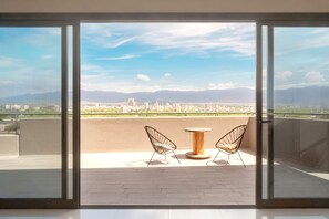 A balcony with two wireframe chairs and a round wooden table, overlooking a cityscape with mountains in the distance.