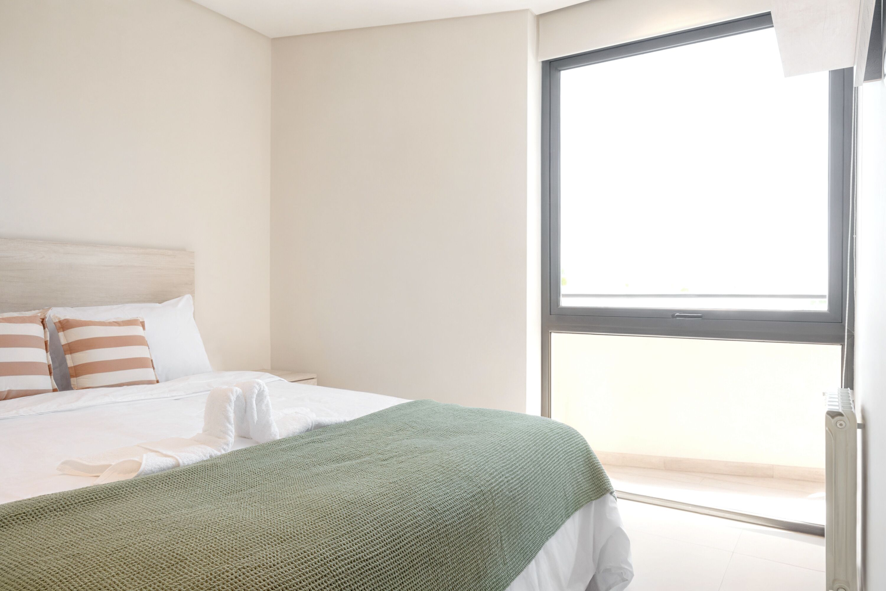 Un dormitorio con cama, almohadas, una manta verde y un ventanal amplio.