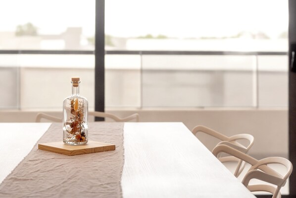 Una mesa con una botella de líquido sobre un posavasos, sillas blancas y vista de un edificio a través de una ventana.