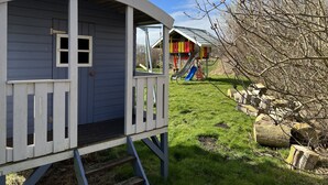 Una casita de juegos de madera pequeña y elevada, con ventana y puerta, junto a un parque infantil colorido con un tobogán.
