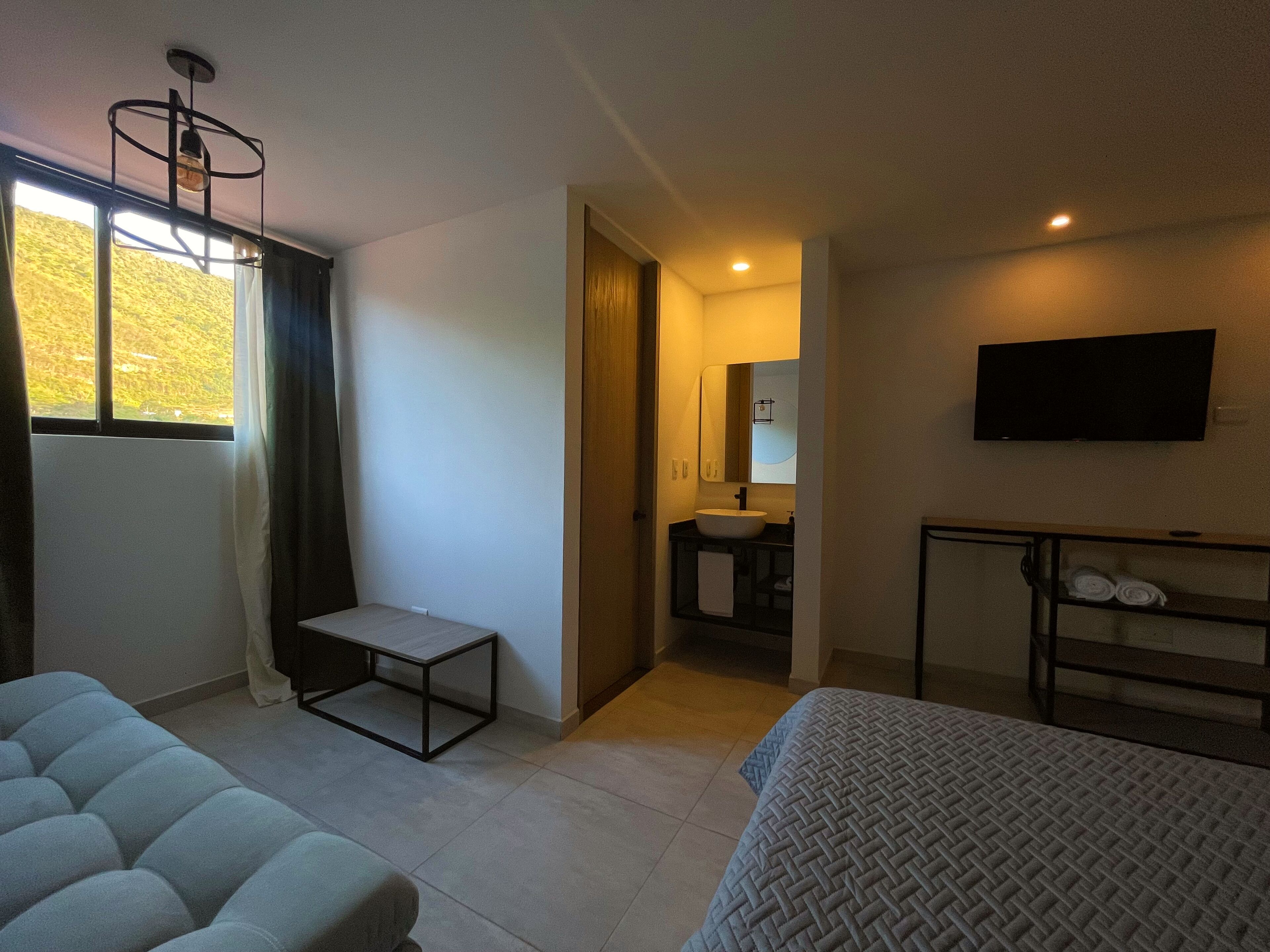 A hotel room with a bed, a small table, a TV, a bathroom with a sink and mirror, and a window with curtains.