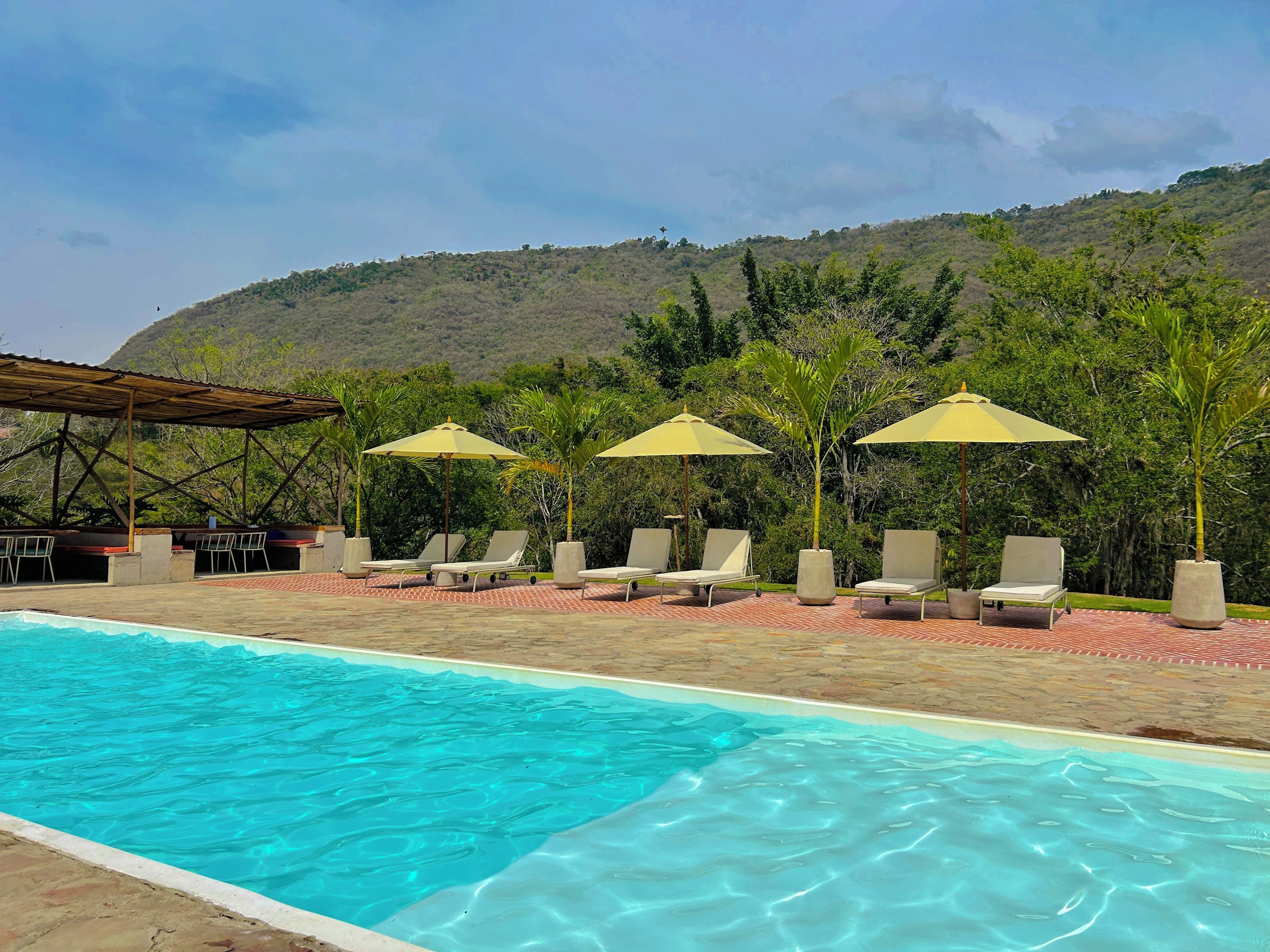 A pool area with lounge chairs and umbrellas, surrounded by greenery and hills.