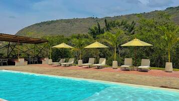 A pool area with lounge chairs and umbrellas, surrounded by greenery and hills.