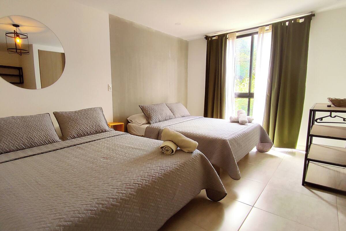A hotel room with two beds, a round mirror, a small table, and a window with curtains.