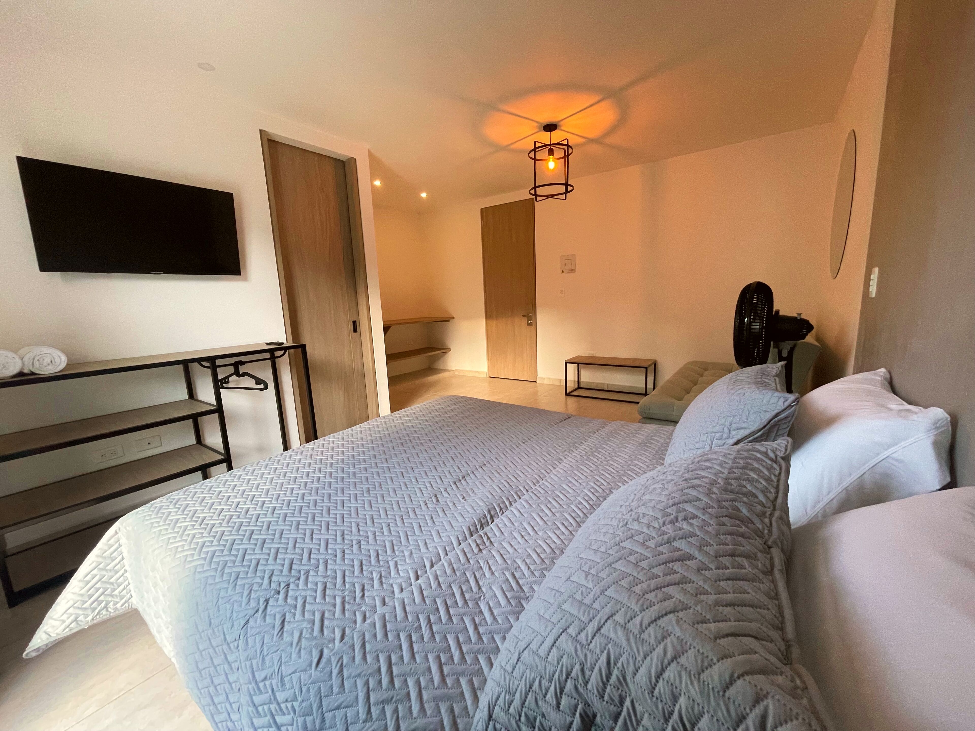 A bedroom with a bed, a TV mounted on the wall, a wooden door, a small table, and a fan.